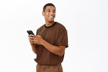 Mobile technology concept. Smiling african american man using his smartphone, holding phone and looking happy, texting in application, white background