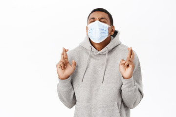 Covid awareness, healthcare concept. African american man in medical mask cross fingers, praying, making wish, hoping for smth, white background