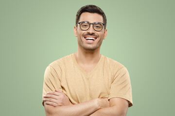 Young male in beige t-shirt and eyewear with crossed arms laughing at joke on green background