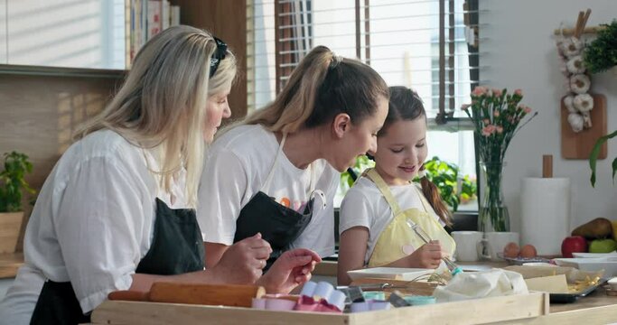 Happy delighted family standing at table in modern kitchen wearing aprons. Young mother with dranny teaching preschooler daughter making homemade domestic cookies biscuits applying egg yolk on it.