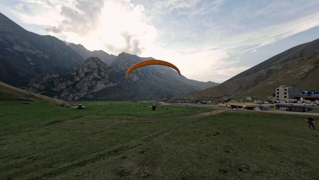 FPV sports drone shot paraglider landing on countryside camp field at mountain valley extreme leisure activity. Aerial view paraplane flying at cliff meadow adventure achievement nature landscape 4k