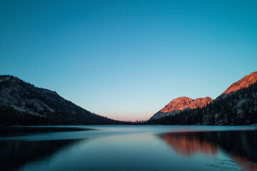 Toxaway Lake, located in Idaho’s Sawtooth Wilderness seen on a summer evening at sunset, with a mountain ridge illuminated reddish orange with the last rays of sunlight. The water reflects the scene.