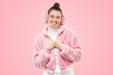 Young smiling girl in pink furry jacket and earmuffs, holding hands close to chest, feeling shy