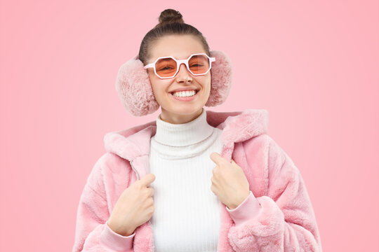 Young trendy teen girl wearing pink fur coat, earmuffs and colored glasses, smiling happily