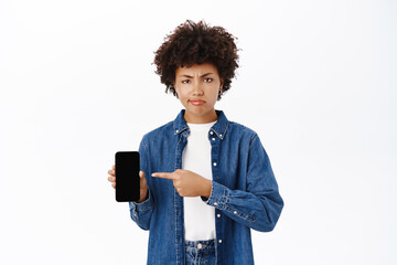 Upset young woman points at her mobile phone and frowns, shows disappointing message or notification on smartphone, white background