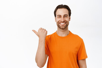 Fototapeta premium Close up of enthusiastic adult man pointing left, smiling amazed, showing advertisement aside, wearing orange t-shirt, white background