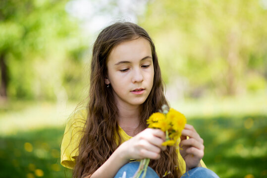 A cute girl on a walk. It's spring outside and the sun is bright. A girl holds dandelions in her hands. She has loose long wavy brown hair. She is dressed in a yellow t-shirt. Childhood. Beauty.