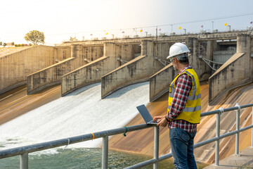 Aืn Engineer standing by the dam. He is wearing a white hard hat and yellow transparent vest.