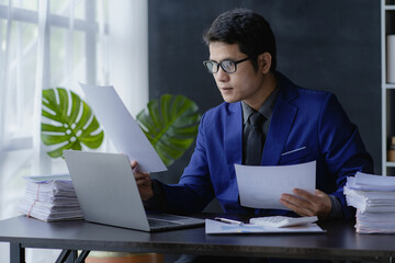 Young financial market analyst wearing glasses works in the office with his laptop while sitting at a wooden table. Asian businessman analyzing documents in hand with document graph