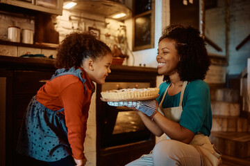 Happy African American girl smells freshly baked sweet pie made by her mother in kitchen.