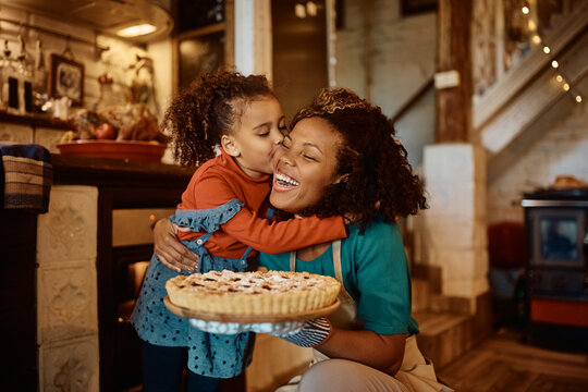 African American mother getting kiss from her daughter after baking sweet pie in kitchen.