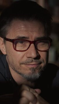 Portrait of good looking man at home in a dark room putting on glasses. Looking away first than looking up and away and smiling 4K video footage.