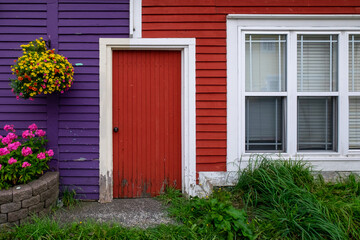 A purple exterior clapboard wooden wall with a hanging flower basket. There's a red door joining the purple wall to a red clapboard wall with a double hung window. There are a flower box and grass too