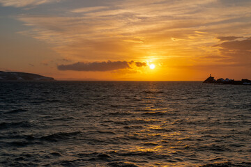 A bright orange and red sunset over the ocean with wispy clouds. The sky has some dark clouds and the sun is just going down over the horizon. There are ripples in the blue water in the foreground. 