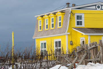 A row of yellow colored window dormers with white frames. The roof is made of cedar shakes from a vintage house. There's a twig fence in the yard in front of the building with a blue sky in the back.