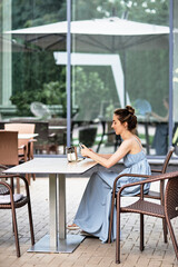 Ukraine. Kharkiv. August. 2022. Photo of a girl with a bun on her head, sitting at a table in a city street cafe, she holds a phone in her hands, the windows of the building are sealed with yellow tap