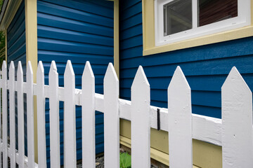 A single hung window in a dark blue exterior house wall of a building. It has narrow v-grove vinyl on the wall. The window has a yellow sill. There's a white picket fence in the foreground.