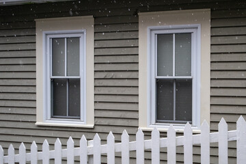 White picket fence on a diagonal in the foreground with a tan colored house in the background. There are two white double hung windows with beige trim on the exterior wall of the country style home. 