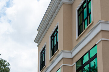 The exterior of a pale yellow colored wall of a house with white decorative trim and molding. The building has multiple windows with green reflective film. There's white clouds in the background. 