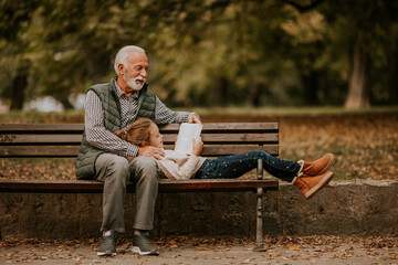 Grandfather spending time with his granddaughter on bench in park on autumn day - obrazy, fototapety, plakaty
