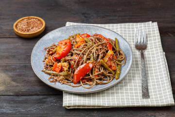 Soba with vegetables in soy sauce sprinkled with flax seeds in a plate on a napkin on a wooden table next to flax seeds in a saucer.