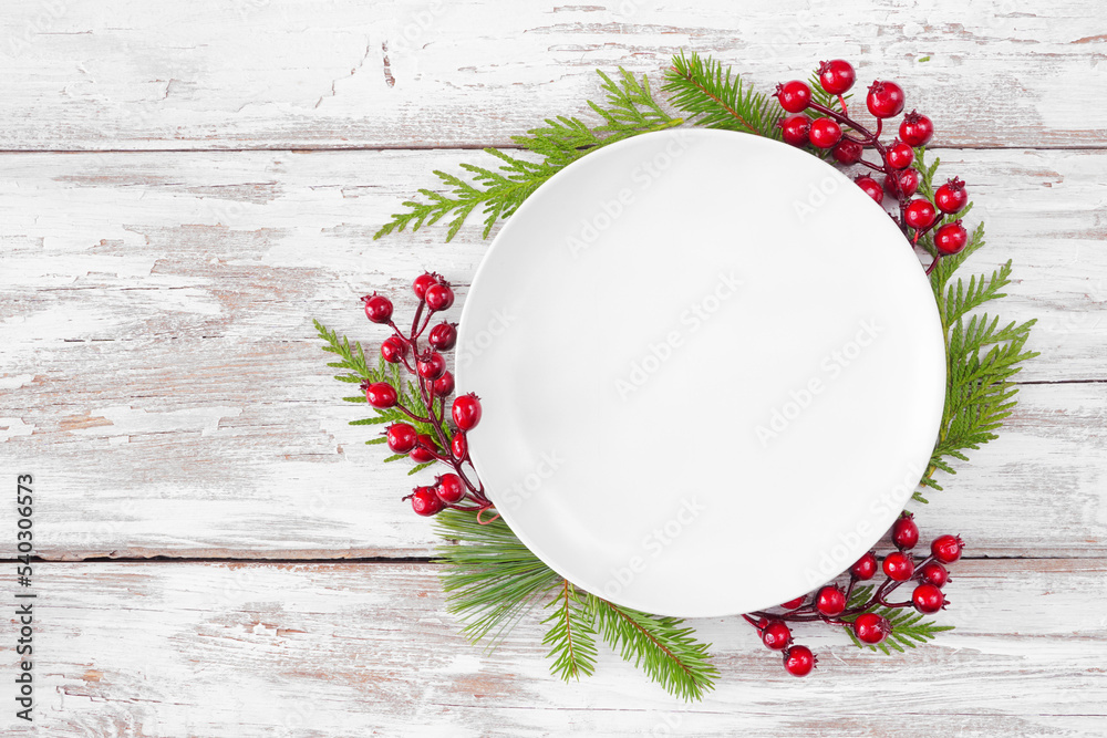 Poster Christmas or winter table setting mock up. Blank white plate with frame of evergreen branches and red berries. Above view on a white wood background. Copy space.