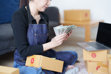 Starting small business entrepreneur of independent Asian woman smiling and holding money using laptop computer with cheerful success of online marketing package box items and SME delivery concept