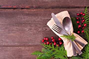 Christmas or winter table setting corner border. Cutlery and napkin  with evergreen branches and red berries. Overhead view on a rustic wood background. Copy space.