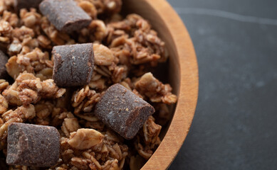Side view of chocolate chunk granola in a wood bowl on a black tile tabletop.