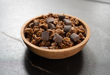 Side view of a small wood bowl filled with chocolate chunk granola on a black tile tabletop.