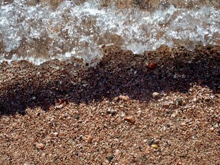 An aerial shot of a wave crashing against a light brown pebbly beach