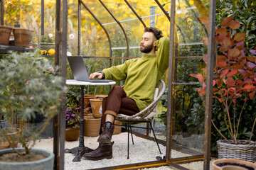 Man works on laptop while sitting by the round table in glasshouse with plants and flowers at...