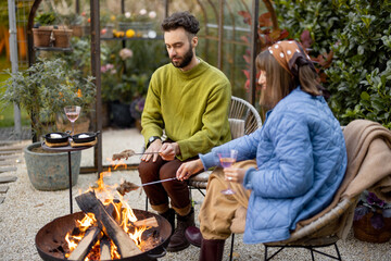 Young stylish couple grilling food and warming up while sitting together by the fire, spending autumn evening time at cozy atmosphere in garden