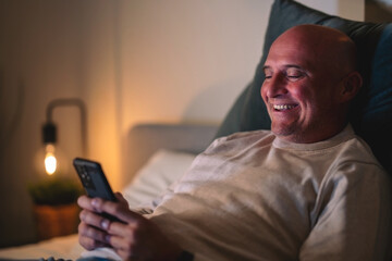 A man is lying on the bed in the room, smiling and reading something good on the phone