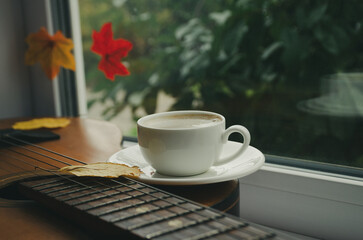 Closeup photo of a coffee cup with fragrant hot coffee, a guitar and autumn leaves.