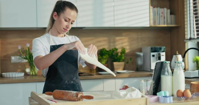 Concentrated serious mother in apron preparing homemade dough using rolling pin cooking baking tasty delicious pizza pasta gnocchi cookies biscuits at home in modern kitchen watching movie on tablet .