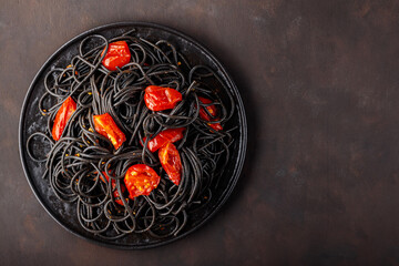 Black spaghetti with cherry tomatoes on a black plate. Boiled black spaghetti pasta with red tomatoes on dark background. Top view. Copy space