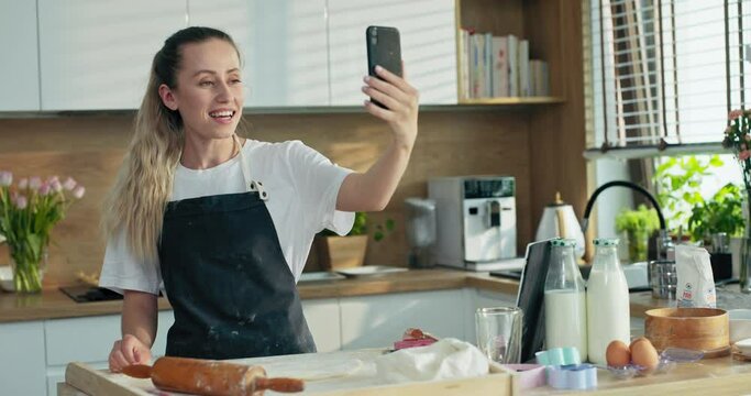 Delighted smiled young mother rolling homemade dough using rolling pin preparing cookies biscuits pizza pasta in modern kitchen talking speaking by video llink with daughter and son.