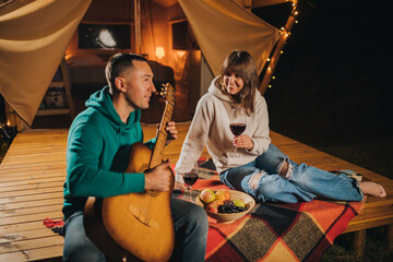 Happy couple relaxing in glamping on autumn evening, drinking wine and playing guitar near cozy bonfire. Luxury camping tent for outdoor recreation and recreation. Lifestyle concept