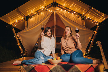 Obraz na płótnie Canvas Two Smiling female friends drinking wine and eating fruits sitting in cozy glamping tent in autumn evening bonfire. Luxury camping tent for outdoor holiday and vacation. Lifestyle concept