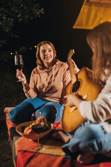 Two Smiling female friends drinking wine and play guitar sitting in cozy glamping tent in autumn evening bonfire. Luxury camping tent for outdoor holiday and vacation. Lifestyle concept