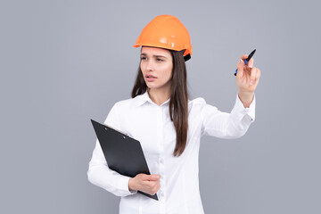 Architect woman in helmet pointing. Young woman in helmet hold clipboard on gray background.