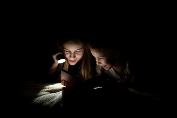 A teenage girl with her sister read in the dark with a flashlight.