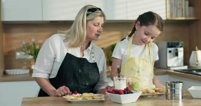 Happy reunion concept. Delighted elderly blonde granny with adorable preschooler granddaughter cooking baking homemade tasty cookies in diferrent shapes decorating with berries. Women making surprise.