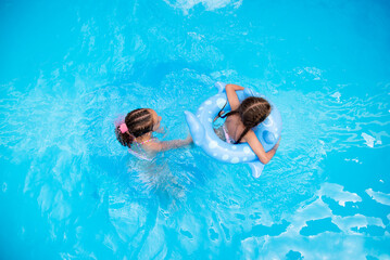 Obraz na płótnie Canvas Two sister girls of 11-13 and 6 years old swim in a pool with blue water and have a fan. The older girl has african braids braided with zi-zi ribbons. Summer. Family vacation.