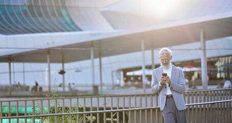 Older mature adult business man, senior middle aged old businessman professional holding cell phone using smartphone mobile apps texting message, working on mobile tech standing in urban city outdoor.