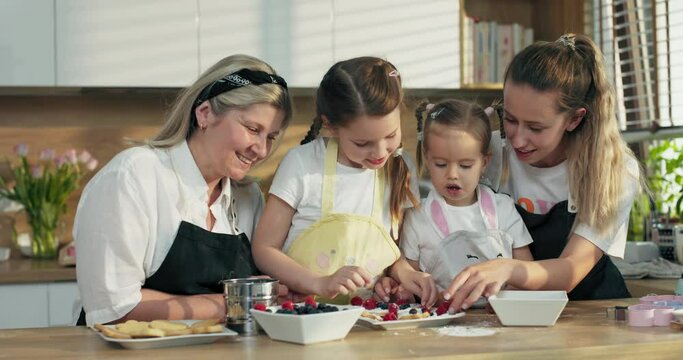 Happy mother's day. delighted middle aged grandmother with daughter and adorable preschooler granddaughters dressing homemade cookies with berries standing at table in modern light kitchen in aprons.