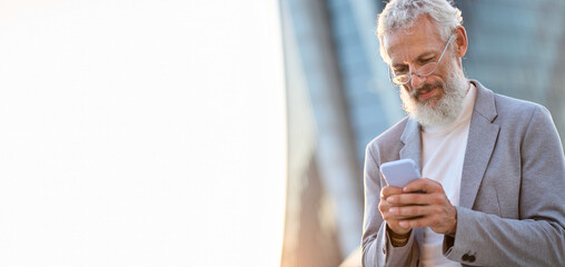 Older middle aged professional business man, smiling senior old businessman wearing suit holding smartphone using mobile cell phone technology standing outdoor in big city office district. Copy space