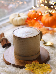 Close up of a cup of healthy pumpkin spices latte with mousse of milk topped with ground spices. Cinnamon sticks and fall leaves with small squash in background. Cozy autumn ambiance, lighting bokeh