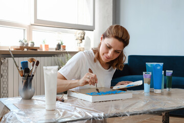 Woman artist painting picture at home with acrylic paints and spatula over the window. Adult hobby and free time creative activity. Female at her 40s enjoying art. 